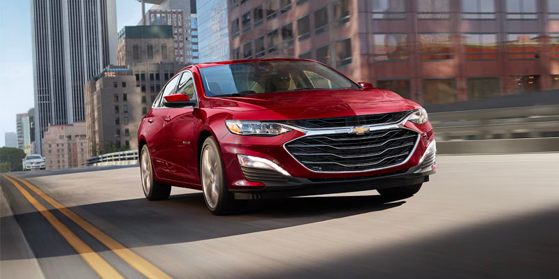 A red 2025 Chevy Malibu driving at speed through downtown Crosby, TX near Turner Chevrolet during daylight. A white car follows in the distant background, with a tall building visible in the right foreground.